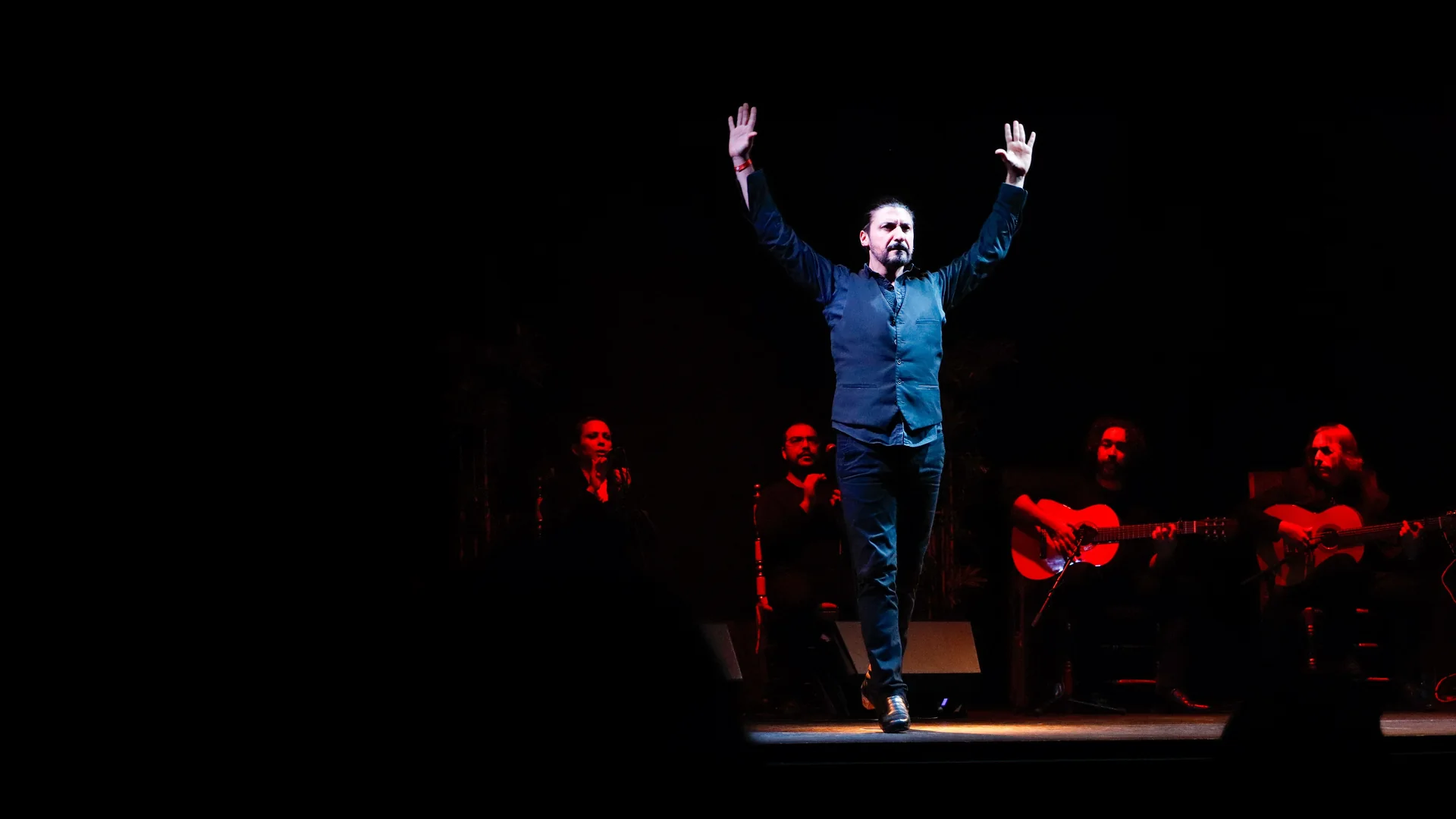 Flamenkový tanečník v tmavě modré vestě stojí se zdviženýma rukama pod jediným reflektorem, za ním na tmavém jevišti sedí zpěvák tleskající do rytmu a dva kytaristé