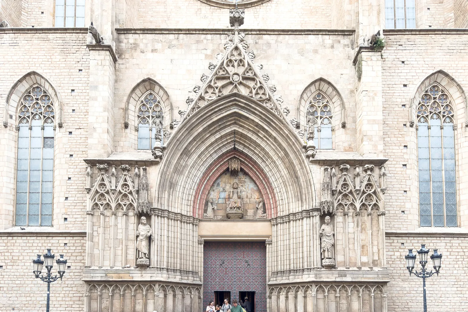 Západní portál Santa Maria del Mar, hrotitý gotický oblouk s vyřezávanými sochami, tmavými dřevěnými dveřmi a návštěvníky stojícími na schodech