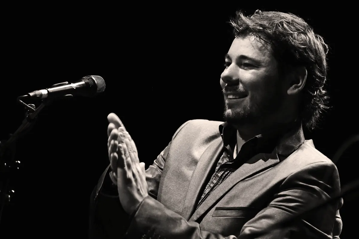 Um cantor de flamenco a sorrir e a bater palmas diante de um microfone num palco escuro